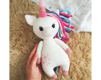 May include: A white crocheted unicorn with a rainbow mane and a pink horn. The unicorn is sitting on a brown fuzzy surface.