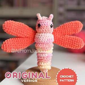 Pode incluir: Uma libélula de crochê com um corpo em tons variados de rosa e amarelo e asas de cor coral sólido. O brinquedo possui olhos de contas pretas e duas pequenas antenas. O texto diz @AllFromJade, ORIGINAL VERSION, CROCHET PATTERN.