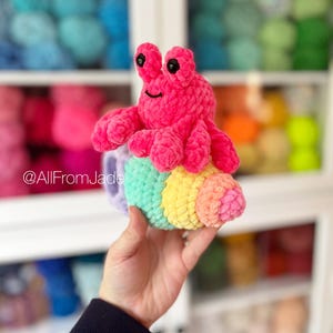 May include: A pink crocheted hermit crab toy with black eyes and a smile, sitting inside a rainbow crocheted shell. The toy is being held in a hand.