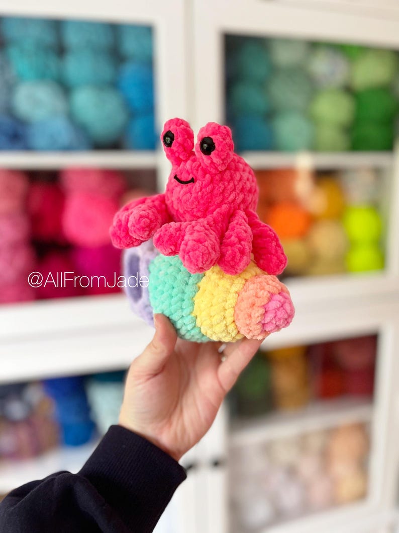 May include: A pink crocheted hermit crab toy with black eyes and a smile, sitting inside a rainbow crocheted shell. The toy is being held in a hand.