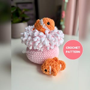 Può includere: Un pesce pagliaccio all'uncinetto fatto a mano con strisce arancioni e bianche, accanto a un anemone di mare all'uncinetto rosa e bianco. Il testo "CROCHET PATTERN" è in un cerchio rosa. È visibile anche il nome dell'artista, "@AllFromJade".