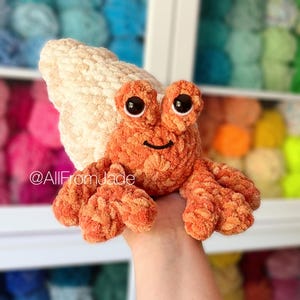 May include: A crocheted hermit crab toy with a white shell and orange body. The crab has large black eyes and a small smile. The toy is being held in a hand.