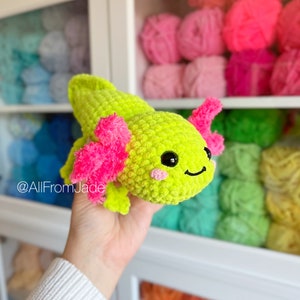 Peut inclure: Un jouet en peluche axolotl crocheté vert vif avec des branchies roses et duveteuses. Le jouet a des yeux noirs et un sourire rose. Le jouet est tenu dans une main sur un fond de pelotes de laine colorées.