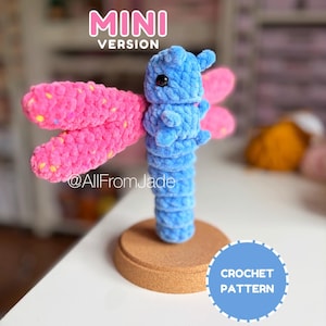 Puede incluir: Un juguete de libélula de peluche con cuerpo azul y alas de color rosa brillante con motas de colores. El juguete tiene ojos de cuentas negras y se asienta sobre una base redonda de corcho. El texto dice MINI VERSION, @AllFromJade y CROCHET PATTERN.