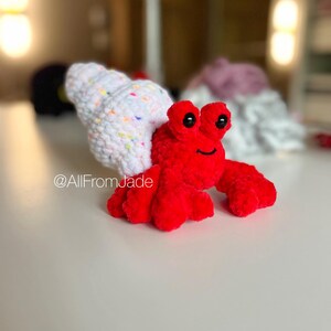 May include: A red crocheted hermit crab toy with a white shell with colorful sprinkles. The crab has black eyes and a smiling mouth. The toy is on a white surface. @AllFromJade