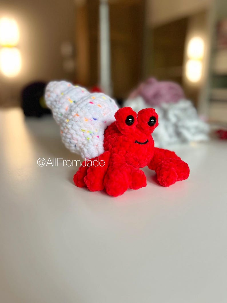 May include: A red crocheted hermit crab toy with a white shell with colorful sprinkles. The crab has black eyes and a smiling mouth. The toy is on a white surface. @AllFromJade