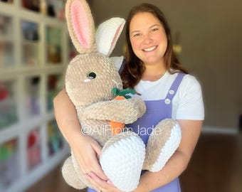 May include: A large, plush, tan-colored bunny with pink inner ears and white accents on the feet. The bunny is holding a small orange carrot with green leaves. The text "@AllFromJade" is visible.