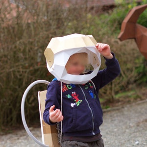 May include: A child wearing a white and gold cardboard astronaut helmet with a blue and white patterned shirt and brown pants. The child is holding the helmet with one hand and has a cardboard rocket pack on their back.