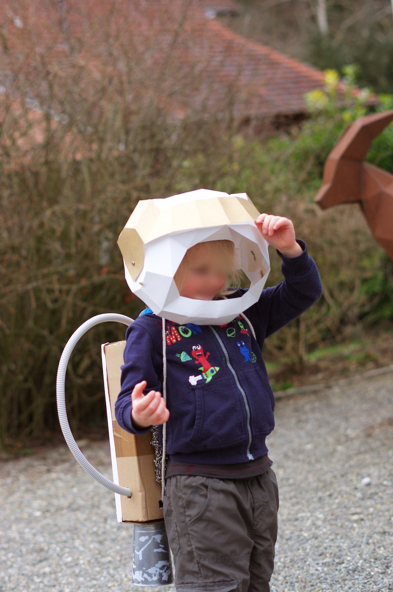 May include: A child wearing a white and gold cardboard astronaut helmet with a blue and white patterned shirt and brown pants. The child is holding the helmet with one hand and has a cardboard rocket pack on their back.