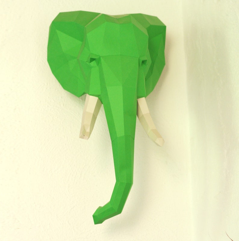 Pu&ograve; includere: Una testa di elefante di carta verde con zanne bianche. La testa di elefante &egrave; un modello 3D ed &egrave; montata su un muro.