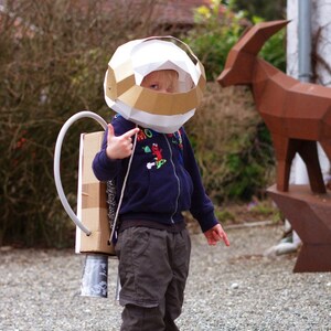 May include: A young child dressed as an astronaut wearing a white and gold cardboard helmet and a blue sweater. The child is holding a cardboard rocket pack with a silver tube attached.
