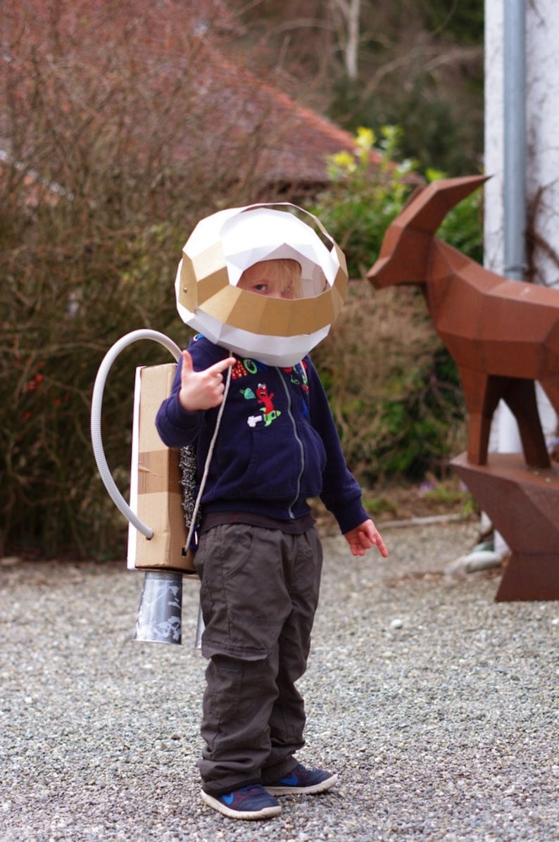 May include: A young child dressed as an astronaut wearing a white and gold cardboard helmet and a blue sweater. The child is holding a cardboard rocket pack with a silver tube attached.