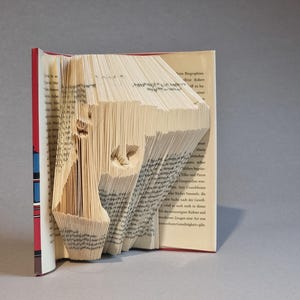 Può includere: Scultura artistica realizzata con un libro. Le pagine sono piegate e tagliate per formare la forma di una pistola. Il libro ha il dorso rosso e le pagine sono beige con testo stampato.