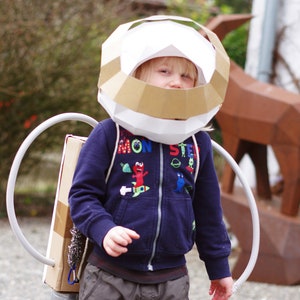 May include: A child wearing a white and brown cardboard astronaut helmet and a blue jacket with a colorful monster design. The child is wearing a cardboard rocket pack with a gray hose.
