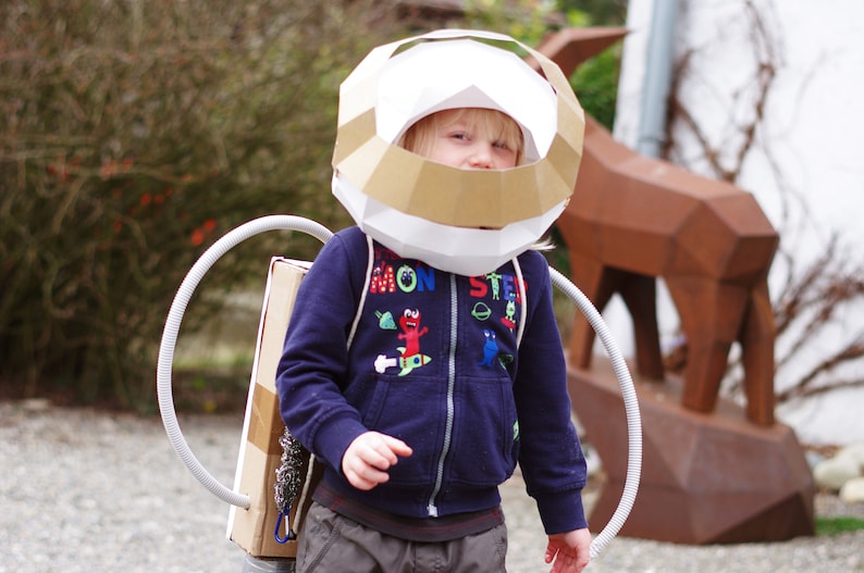 May include: A child wearing a white and brown cardboard astronaut helmet and a blue jacket with a colorful monster design. The child is wearing a cardboard rocket pack with a gray hose.