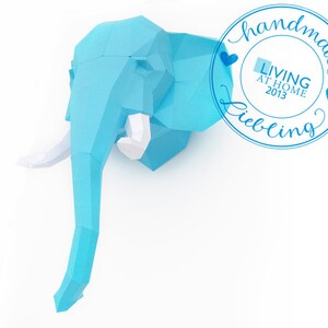 Pu&ograve; includere: Una scultura di testa di elefante in carta di colore azzurro con zanne bianche. La scultura &egrave; fatta a mano e ha un'etichetta "Living at Home 2013" con un design a forma di cuore.