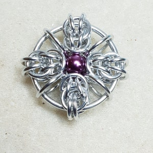May include: A silver metal pendant with a purple bead in the center. The pendant is made of woven metal and has a circular shape.