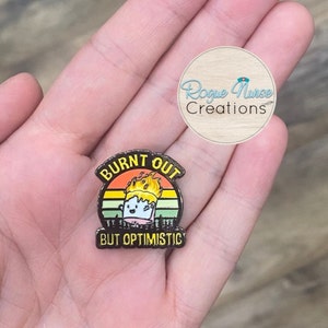 May include: A black enamel pin with the words "BURNT OUT BUT OPTIMISTIC" above and below a cartoon marshmallow on fire. The pin has a sunset background and a forest silhouette.