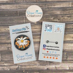May include: A button pin with a black background and a daisy design in white and orange. The pin is on a light blue card with the text "Button Pin" and "Rogue Nurse Creations". The back of the card has a pin clasp.