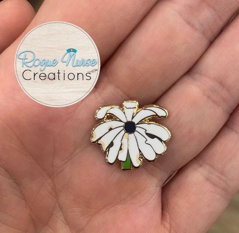 May include: Gold-tone enamel pin featuring a white daisy with a black centre. The pin is on a hand.