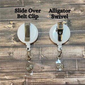 Op de afbeelding: Twee witte intrekbare badge-rollen met verschillende bevestigingen. De rol aan de linkerkant heeft een schuifbevestiging voor een riem. De rol aan de rechterkant heeft een alligator-draaibare clip. Beide rollen hebben transparante plastic badgehouders.