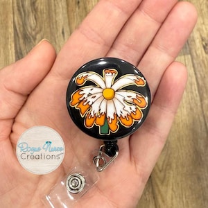 May include: A black retractable badge reel with a daisy design. The daisy has white petals with orange flame-like accents and a yellow center. The reel has a metal clip and a clear plastic snap.