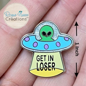 May include: A silver enamel pin featuring a green alien in a blue and purple spaceship with glitter. The spaceship has three purple dots on the bottom. The text "GET IN LOSER" is written in black on a yellow background.
