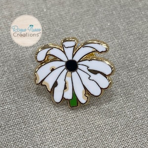 May include: Gold-tone enamel pin featuring a white daisy with a black centre. The daisy has a green stem and is surrounded by a gold outline.