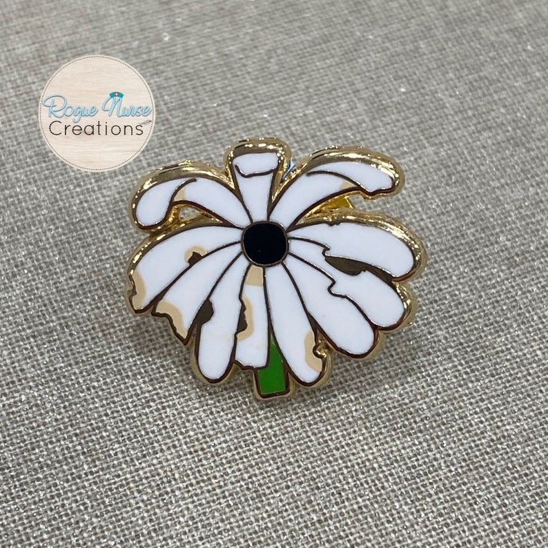 May include: Gold-tone enamel pin featuring a white daisy with a black centre. The daisy has a green stem and is surrounded by a gold outline.