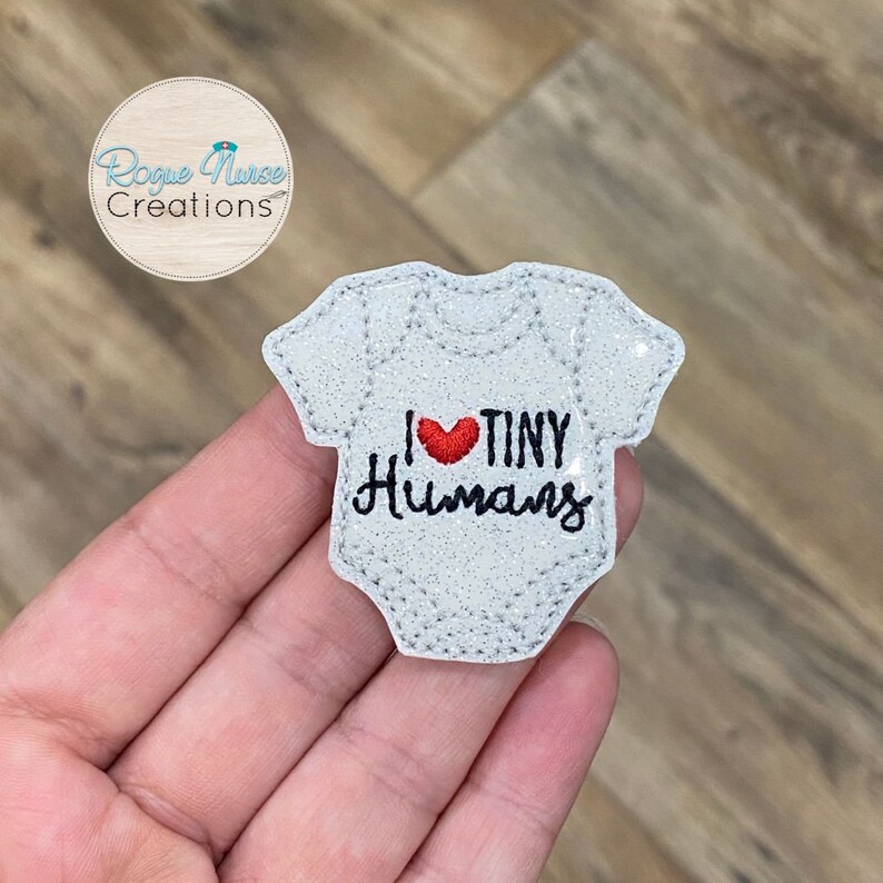 Op de afbeelding: Een witte glitter strijk-op patch in de vorm van een onesie met de tekst "I &hearts; TINY HUMANS" in zwarte borduurwerk en een rood geborduurd hart.
