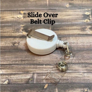 Op de afbeelding: Witte intrekbare badgehouder met een schuifbare riemclip. De houder heeft een transparante plastic band en een zilveren metalen knop.