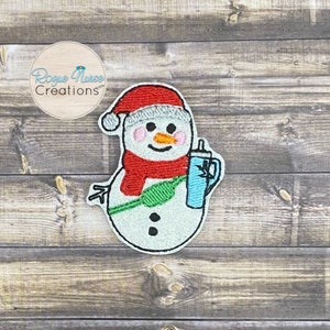 May include: Embroidered snowman wearing a red hat and scarf, holding a blue cup with a straw. The snowman has a green scarf around its neck and two black buttons.