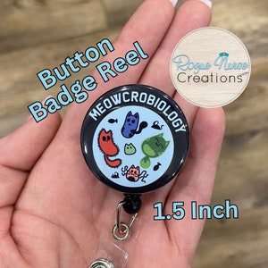 May include: A black button badge reel with a 1.5 inch diameter button featuring a blue background with the text "MEOWCROBIOLOGY" in a black circle and a cartoon illustration of cats.