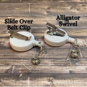Op de afbeelding: Twee witte intrekbare badge-haspels met verschillende clip-stijlen. Een haspel heeft een schuifbevestiging voor de riem en de andere heeft een alligator-draaiclip. Beide haspels hebben transparante plastic banden.