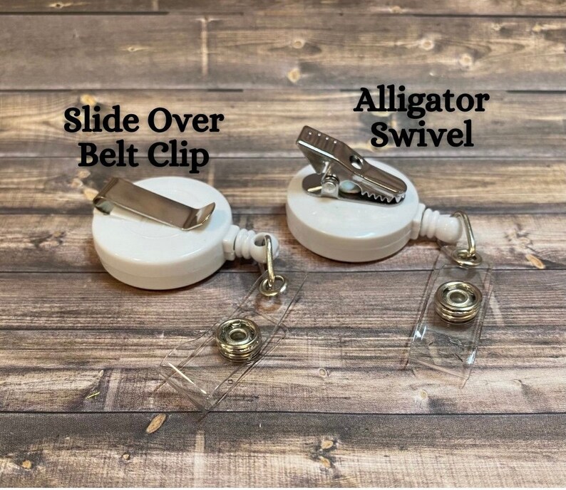 Op de afbeelding: Twee witte intrekbare badge-haspels met verschillende clip-stijlen. Een haspel heeft een schuifbevestiging voor de riem en de andere heeft een alligator-draaiclip. Beide haspels hebben transparante plastic banden.