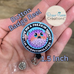 May include: A blue and purple pufferfish with a distressed expression on a 1.5 inch button badge reel. The text on the badge reads "Give me a freakin minute. I'm overstimulated."