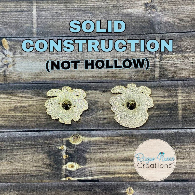 May include: Two gold-coloured enamel pins with glitter detail. The pins are shaped like a flower with a single rhinestone in the centre. The text "SOLID CONSTRUCTION (NOT HOLLOW)" is displayed above the pins. The text "Rogue Nurse Creations" is displayed below the pins.