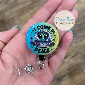 May include: A green alien wearing a flower crown and holding a peace sign is on a colorful badge reel with the text "I Come In Peace".