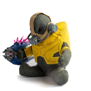 Op de afbeelding: Een pluche speelgoed in een gele en grijze beschermende outfit met een bruine helm en een gasmasker. Het speelgoed houdt een blauw en roze klauwachtig wapen vast. Het speelgoed zit op een witte achtergrond.