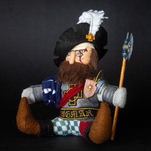 May include: A plush toy figure of a warrior wearing a black hat with a white feather, a grey and blue tunic, and a silver breastplate with a red sash and a gold cross. The figure is holding a wooden staff with a silver axe head. The figure has a brown beard and a bandage over one eye. The figure's name, "Sigmar", is written on the breastplate.