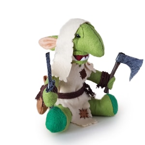 Puede incluir: Un juguete de peluche de goblin verde con una capucha blanca, sosteniendo un pequeño hacha y un cuchillo. El goblin lleva un cinturón marrón y una túnica blanca con parches marrones.