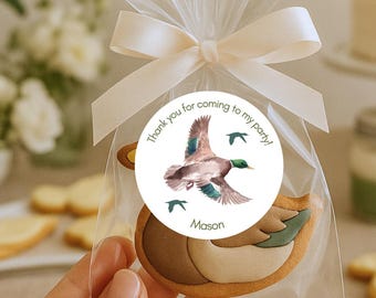 Puede incluir: Una bolsa transparente atada con una cinta crema, que contiene una galleta con forma de pato. Una pegatina redonda en la bolsa dice "Thank you for coming to my party!" con una ilustración de pato en acuarela y el nombre "Mason".