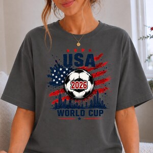 USA Soccer World Cup 2026 graphic shirt featuring soccer ball, American flag splash art, skyline design and patriotic football fan aesthetic