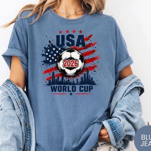 USA Soccer World Cup 2026 graphic shirt featuring soccer ball, American flag splash art, skyline design and patriotic football fan aesthetic