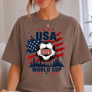 USA Soccer World Cup 2026 graphic shirt featuring soccer ball, American flag splash art, skyline design and patriotic football fan aesthetic