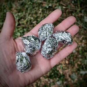 May include: A hand holding five small, irregularly shaped objects wrapped in shiny silver foil. The objects are clustered together, with the background showing out-of-focus green foliage.
