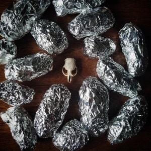 May include: A collection of silver foil-wrapped packages, some with a crinkled texture, arranged on a wooden surface. A small white animal skull is in the center of the arrangement.