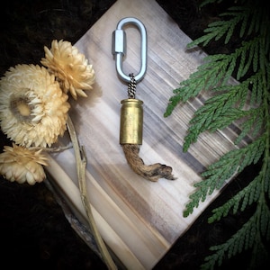 May include: A brass bullet casing with a brown leather tassel attached to a silver key chain. The keychain is on a wooden surface with a green leafy branch.