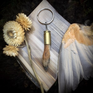 May include: A brass bullet casing keychain with a small animal foot attached. The foot is brown and furry. The keychain is hanging from a silver key ring.