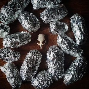 May include: A still-life arrangement featuring a small animal skull surrounded by numerous foil-wrapped objects. The skull is off-white, and the foil has a metallic silver sheen. The items are arranged on a dark brown wooden surface.